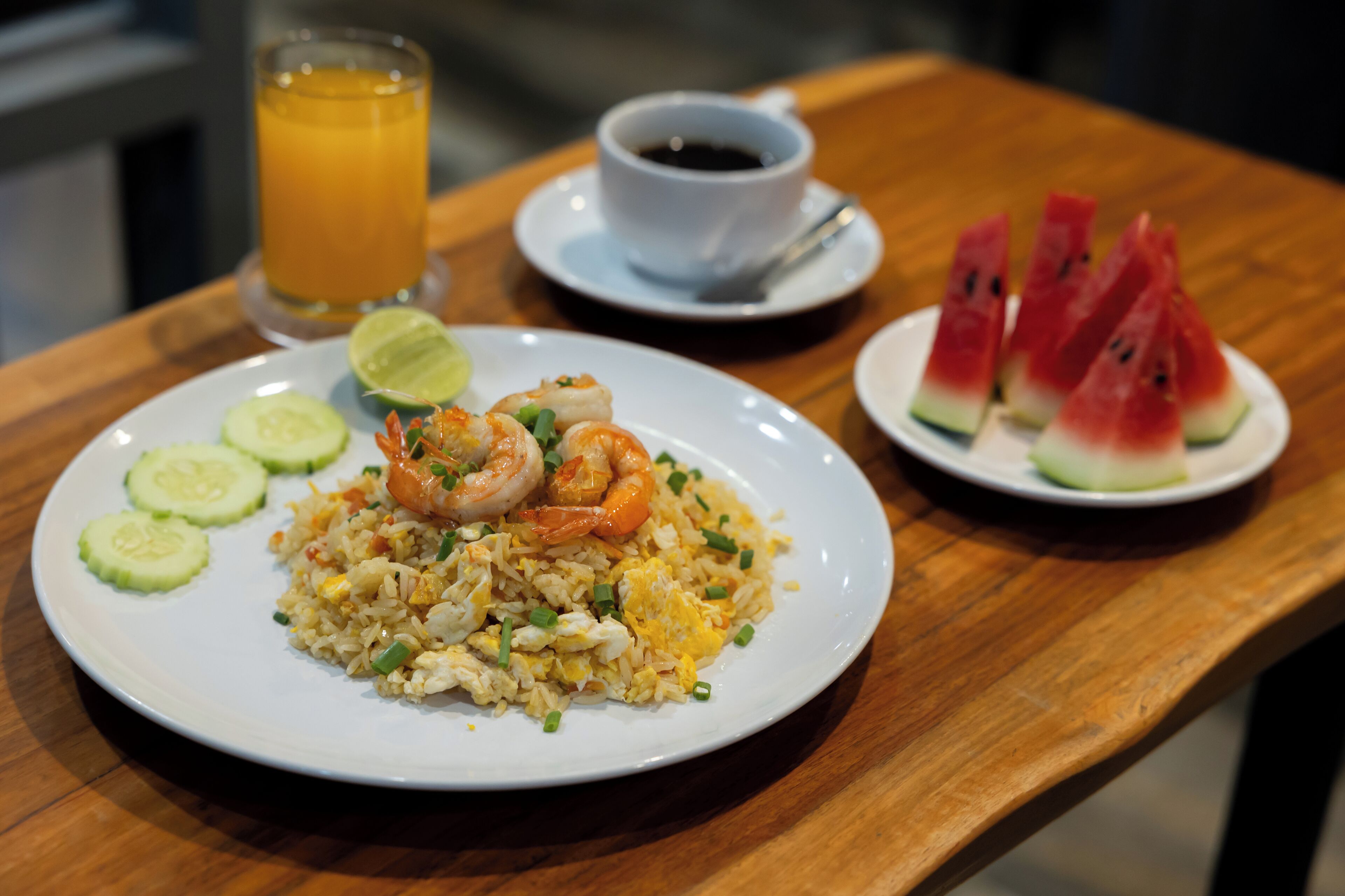 A plate of fried rice with shrimp and cucumber slices, a glass of orange juice, a cup of black tea, and a plate of watermelon slices.