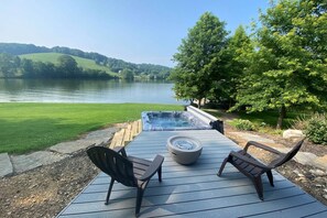Un jacuzzi en una terraza de madera con dos sillas, con vistas a un lago y colinas.