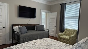 Un dormitorio con cama, televisión en la pared, una silla y ventana con persianas.