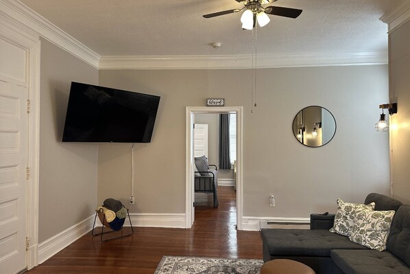 A living room with a flat-screen TV mounted on the wall, a ceiling fan, a large mirror, and a gray sofa with patterned pillows.
