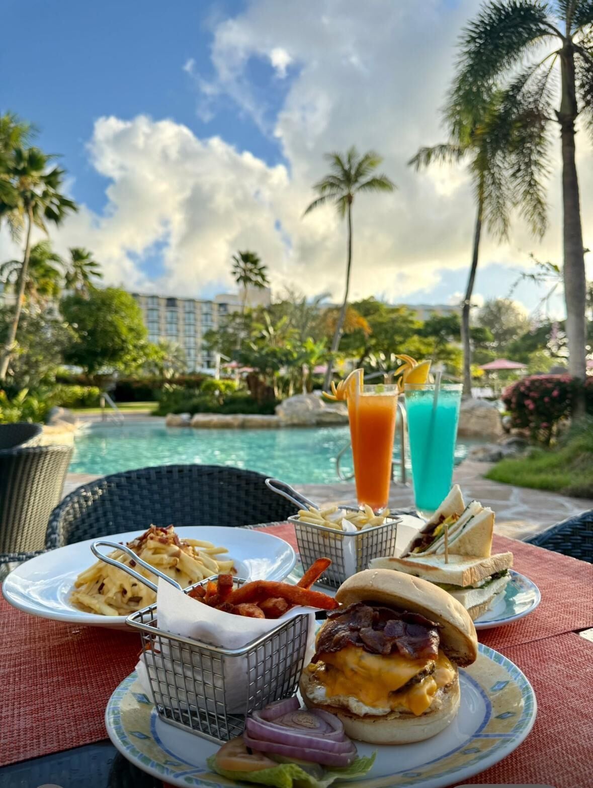 A dining table with a meal consisting of a burger, fries, and a drink, set against a poolside view with palm trees and a building in the background.