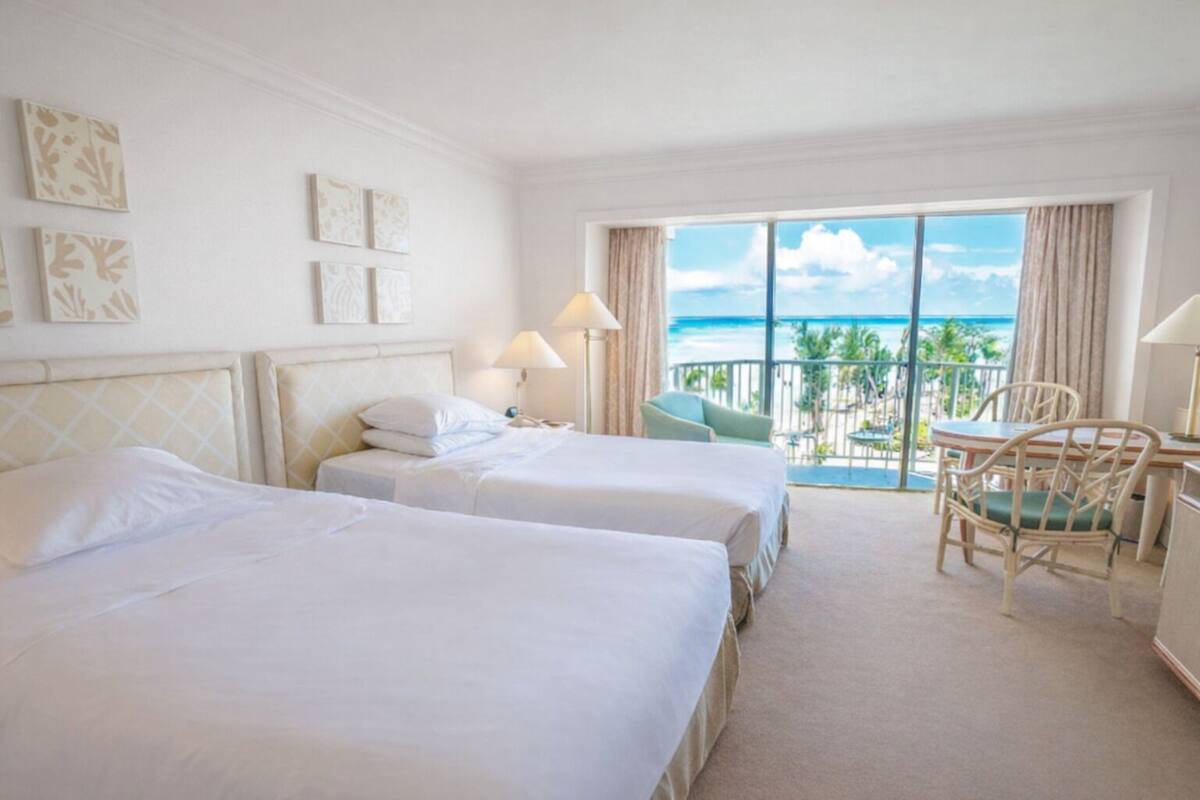 A hotel room with two beds, a balcony with a view of the ocean, and a dining area with chairs.
