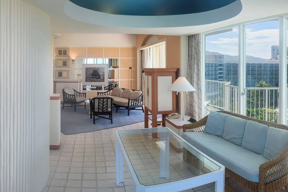A hotel room with a bed, a sofa, a dining area with chairs, and a view of the city.