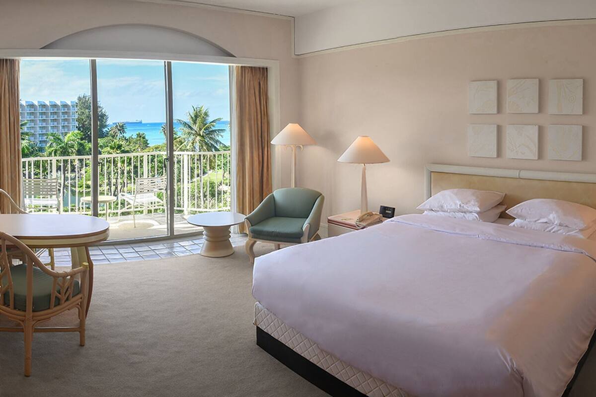 A hotel room with a bed, two bedside tables, a chair, a small table, and a balcony with a view of the ocean and palm trees.