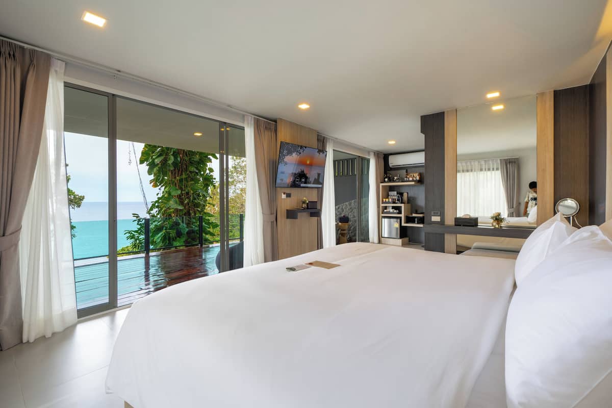 A large bed in a room with a view of the ocean, a small desk with a mirror, and a bathroom with a shower.