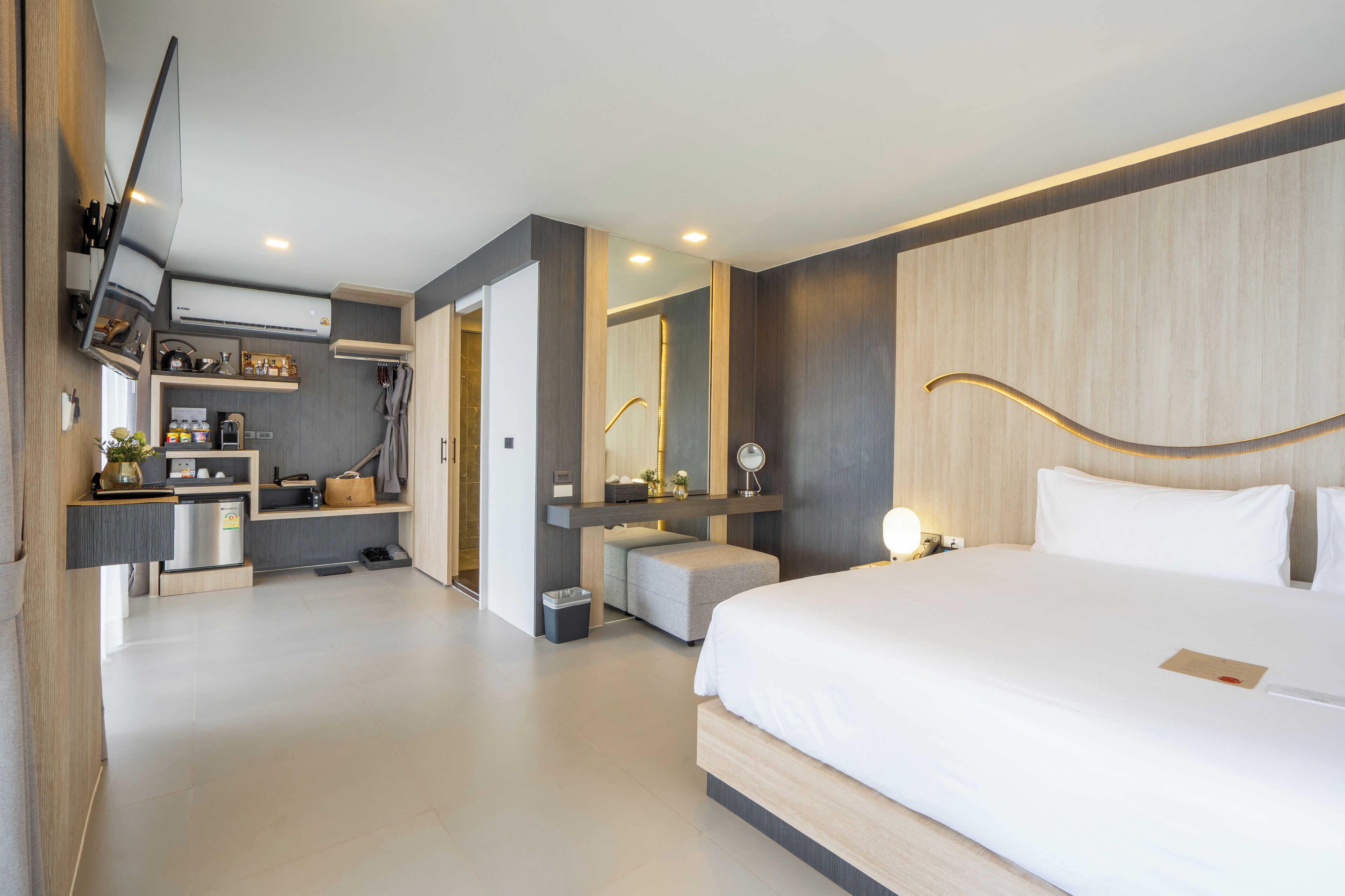 A modern hotel room with a large bed, a built-in desk, a vanity with a mirror, and a kitchenette.