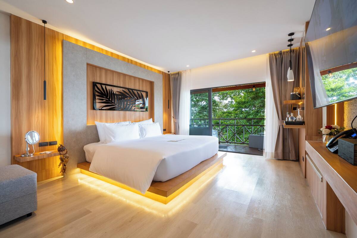 A modern bedroom with a large bed, a bedside table with a lamp, a small seating area, and a balcony with a view of greenery.