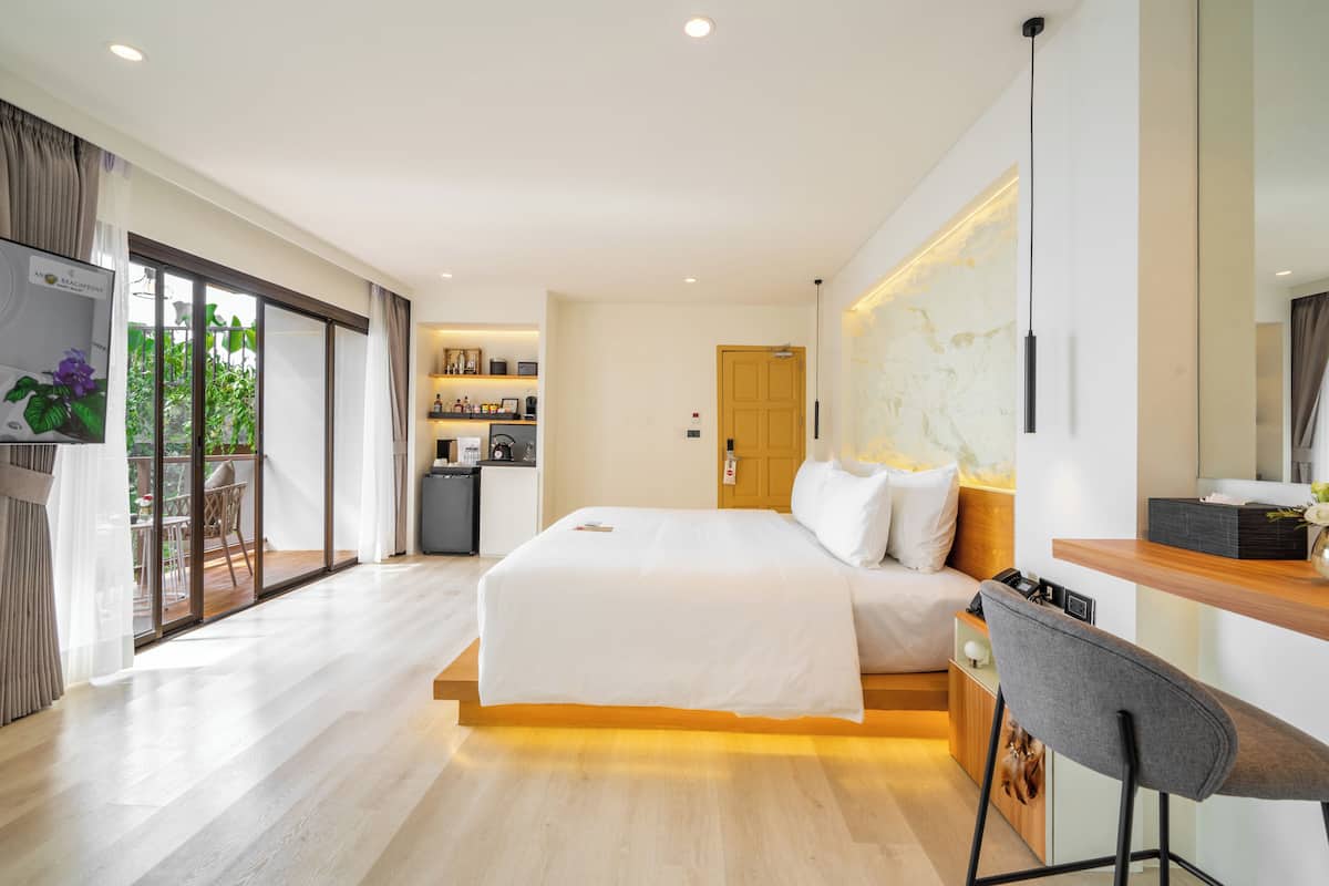 A modern hotel room with a large bed, a desk with a chair, a small kitchenette, and a sliding glass door leading to a balcony.