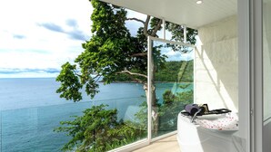 A balcony with a glass railing overlooking a coastal view, a white bathtub, and a small seating area with towels. - Anona Sunset Boutique (Karon)
