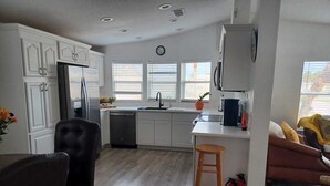 A modern kitchen with white cabinets, a dishwasher, and a sink. There is a wooden stool and a brown chair. A clock is on the wall, and a fire extinguisher is visible. - Find your perfect home in Fort Myers - Affordable, comfortable & ready for you  (Fort Myers)
