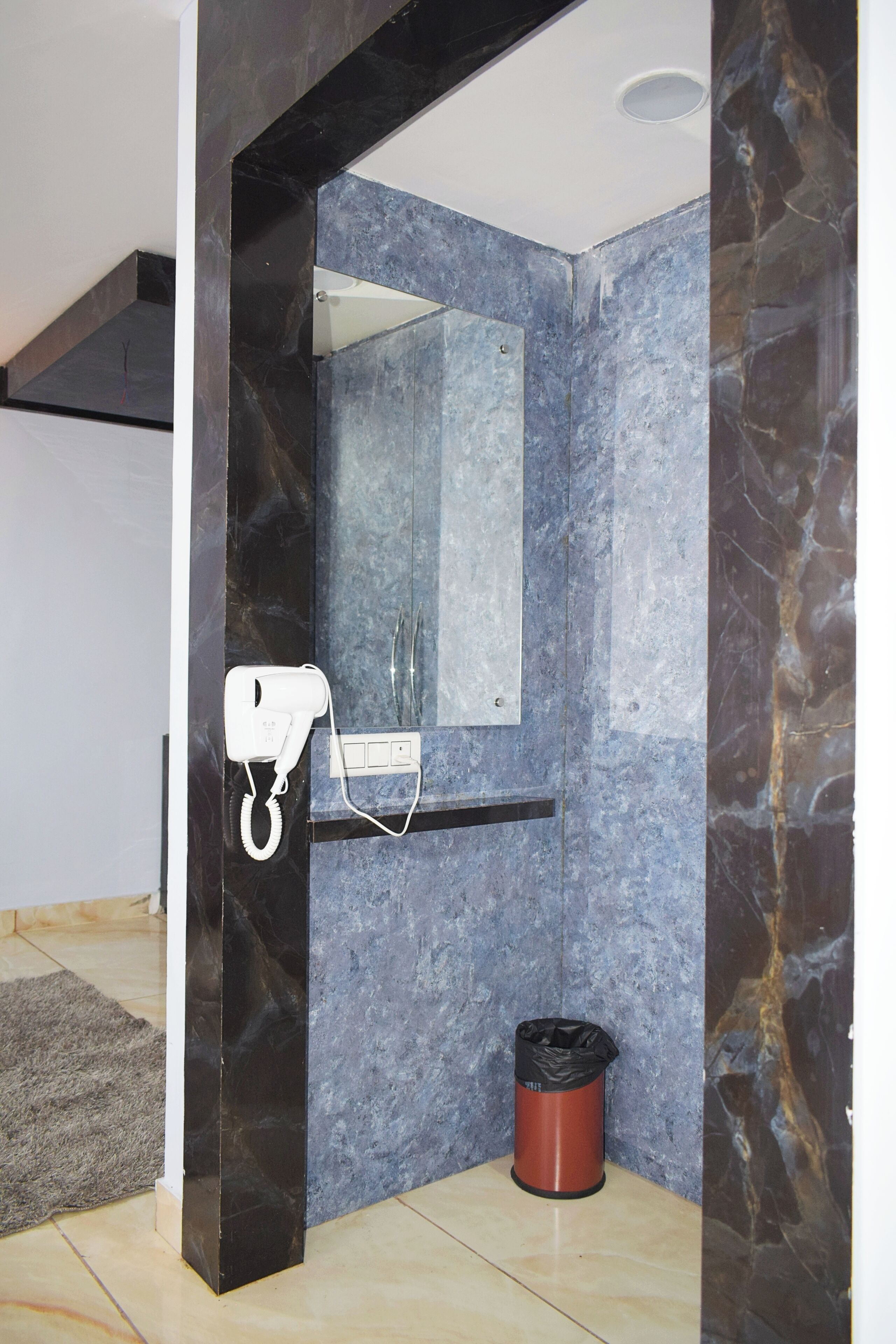 A bathroom with a marble wall, a hairdryer, a mirror, and a trash bin.