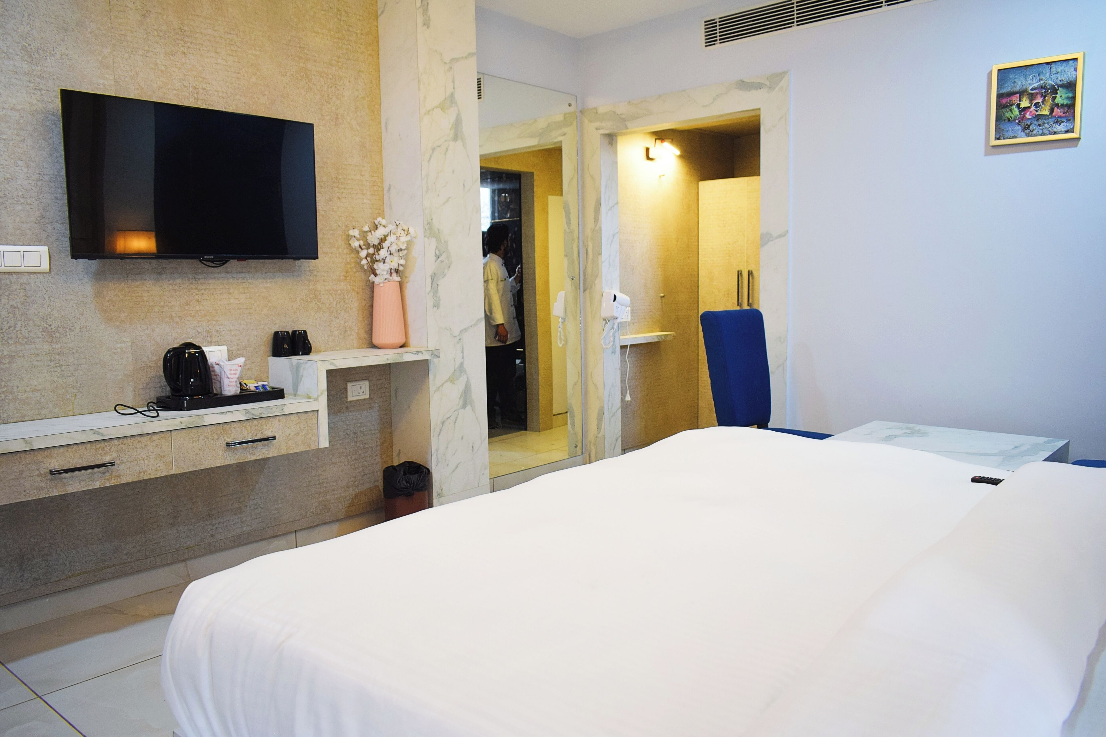 Une chambre d’hôtel avec un lit, un téléviseur fixé au mur, un miroir, un bureau avec un téléphone et une salle de bain visible à travers une porte ouverte.