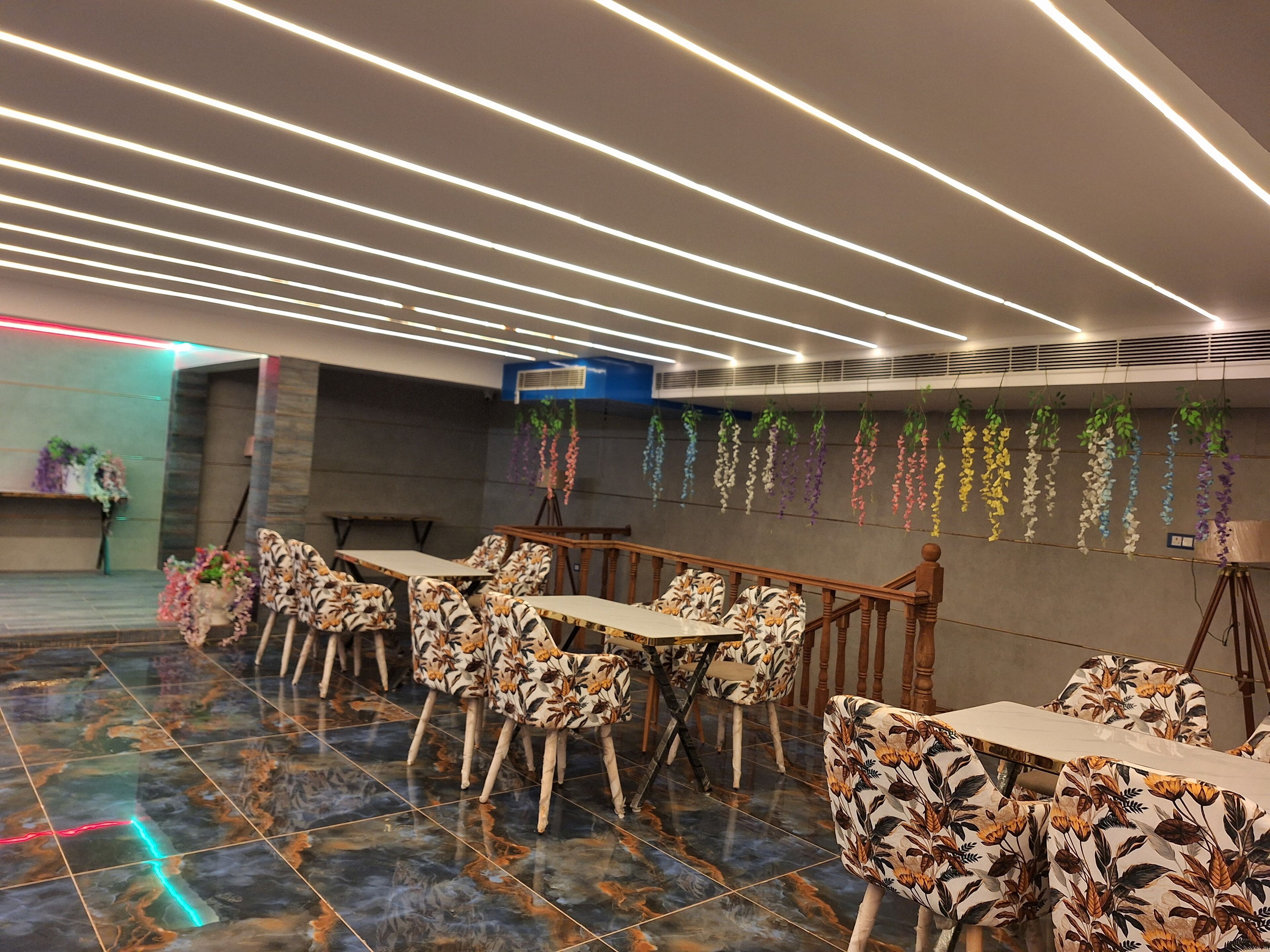 Un espace de salle à manger moderne, avec une longue table et des chaises, un plafond doté d’un éclairage linéaire et des plantes suspendues.