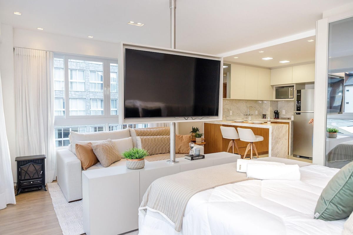 A modern living room with a large flat-screen TV, a sofa, a bed, and a kitchen area with a dining table and chairs.