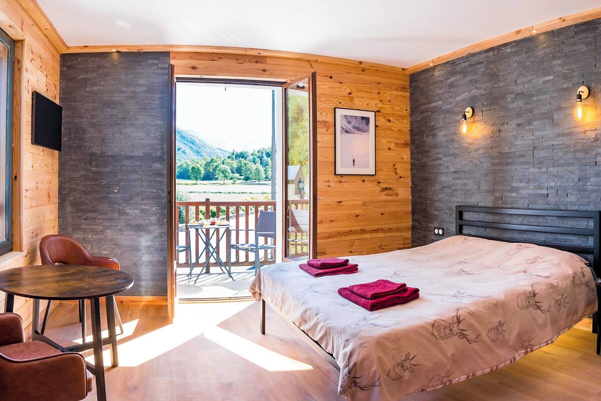 A bedroom with a bed, a small table, and chairs, a wooden wall, and a view of the outdoors.