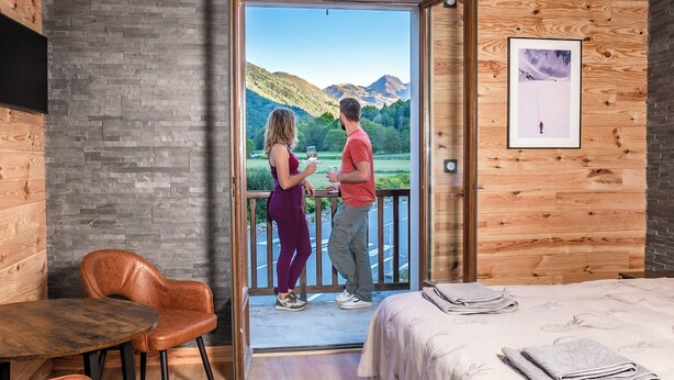A room with a wooden interior, a bed with a white bedspread, a wooden table with a chair, and a view of a mountainous landscape through an open door. - Apart Hotel des Cols  (Borderes-Louron)