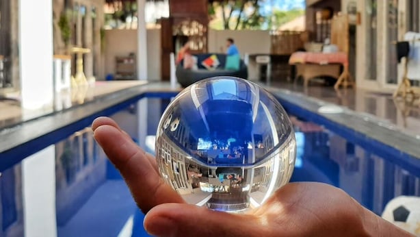 Een hand houdt een glazen bol boven een zwembad, dat de omgeving weerspiegelt.