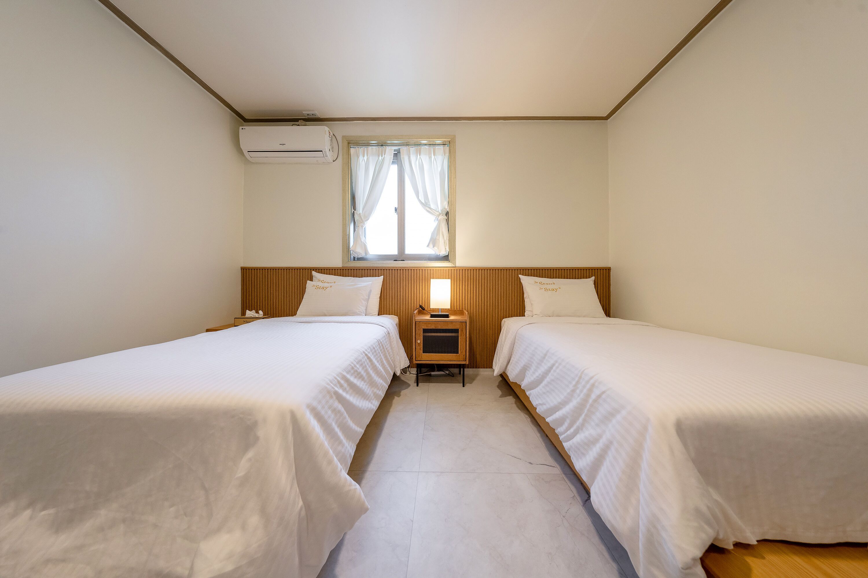 Una habitación con dos camas individuales, una mesita de noche con lámpara, un equipo de aire acondicionado y una ventana con cortinas.
