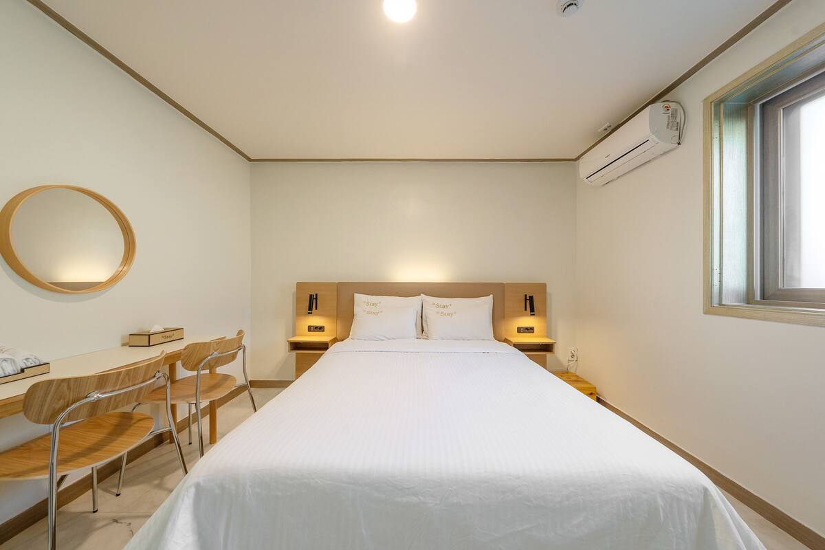 A hotel room with a bed, two bedside tables, a desk with chairs, a round mirror, and an air conditioning unit.