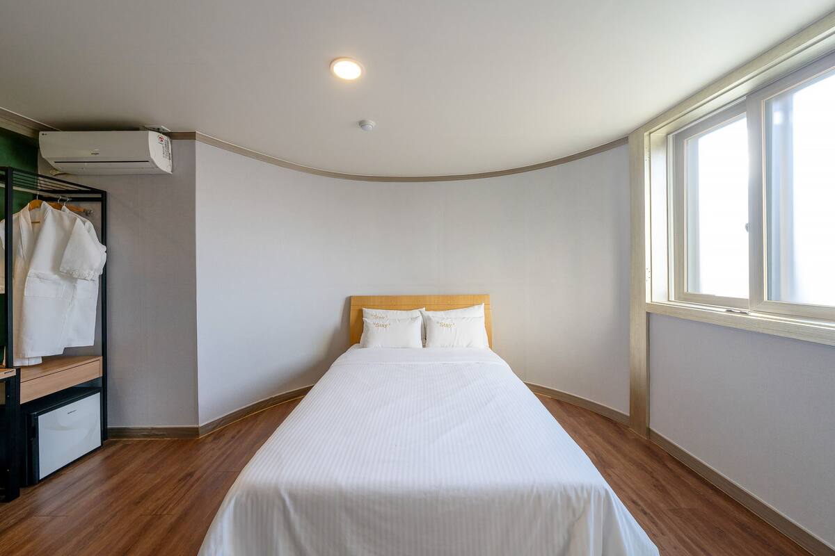 A neatly made bed with a wooden headboard in a room with a wooden floor, a window on the right, and a wall-mounted air conditioner.
