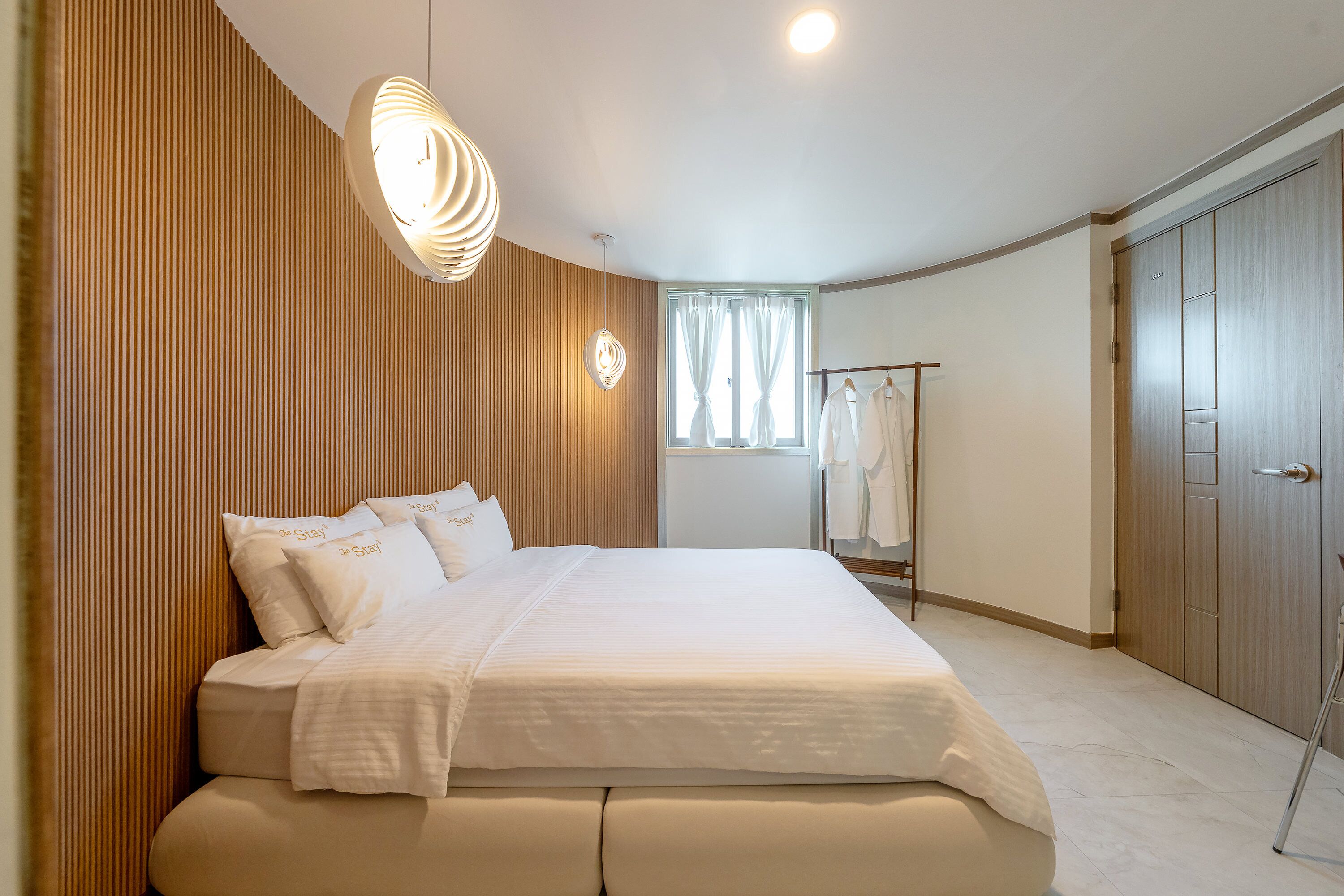 A neatly arranged bedroom with a large bed, a hanging light fixture, a wardrobe, and a window with curtains.