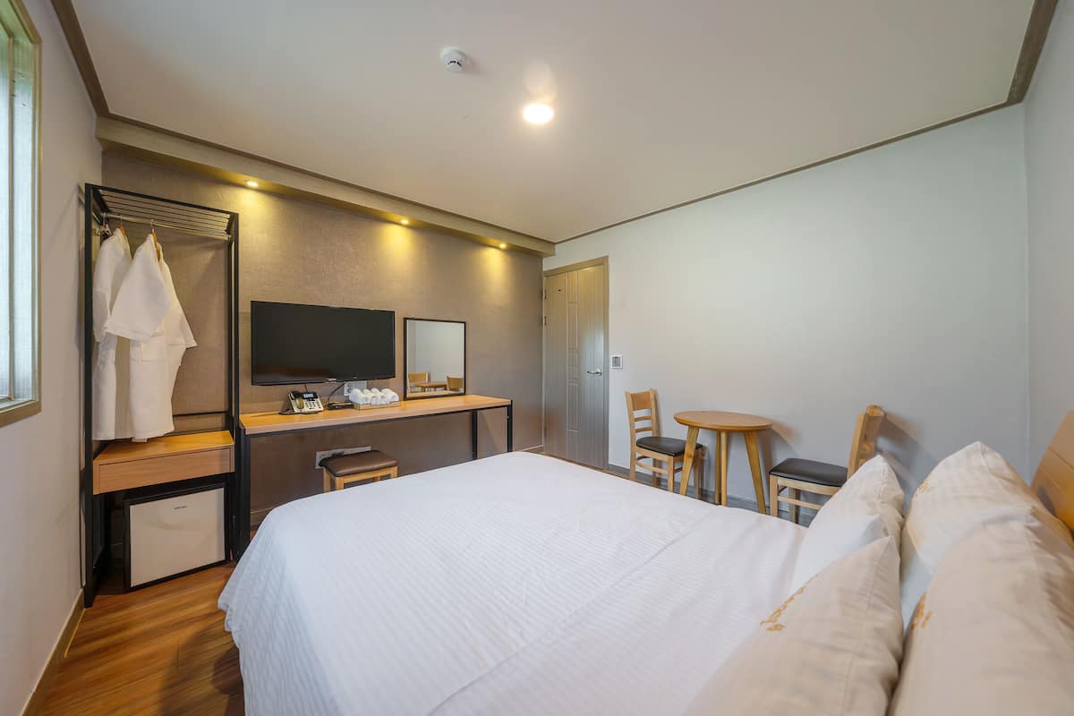 A hotel room with a bed, a desk with a computer, a chair, a small table, a mirror, and a wardrobe.