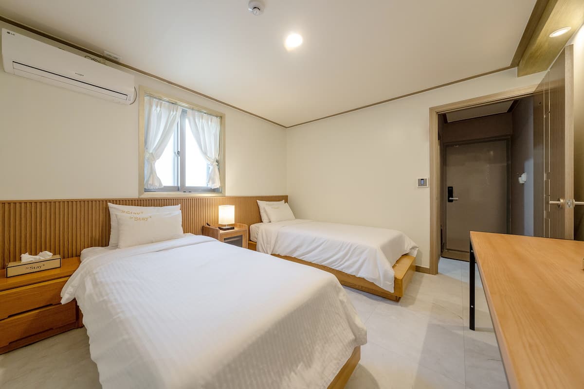 A hotel room with two beds, a wooden headboard, a bedside table with a lamp, a window with curtains, and a door.