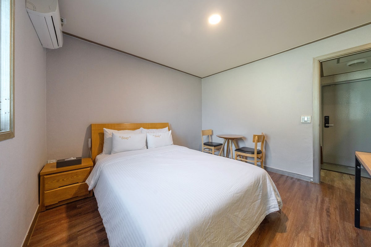 A hotel room with a large bed, wooden nightstand, two chairs, a small table, and a door leading to a bathroom.