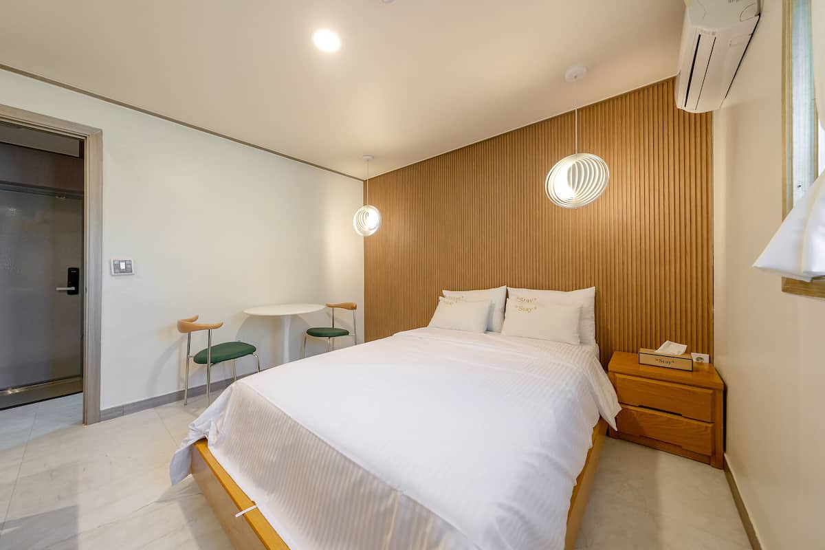 A modern hotel room with a large bed, a bedside table with a phone, a small round table with a chair, and a wall-mounted air conditioner.