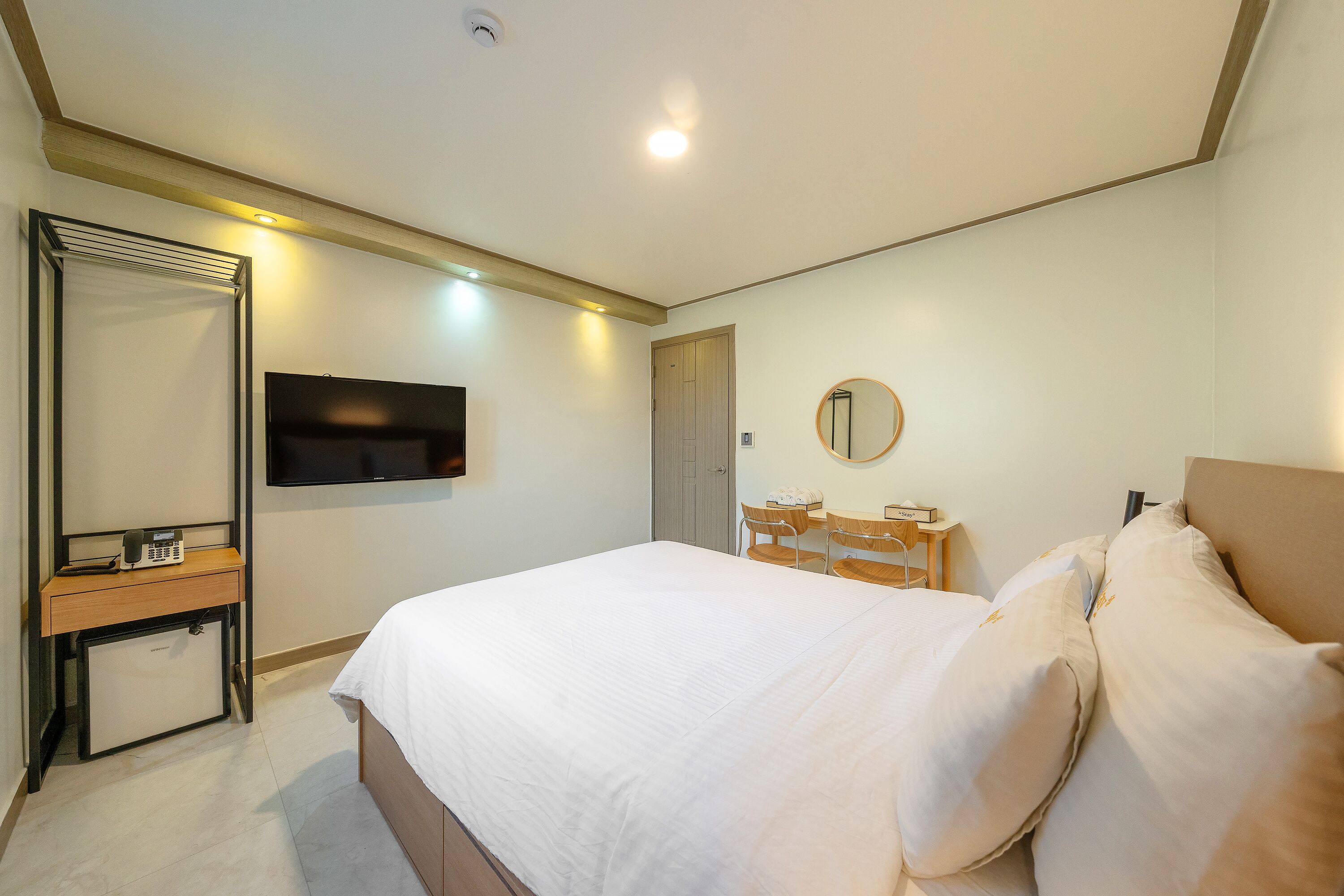 A modern hotel room with a bed, a desk with a chair, a TV, and a mirror.