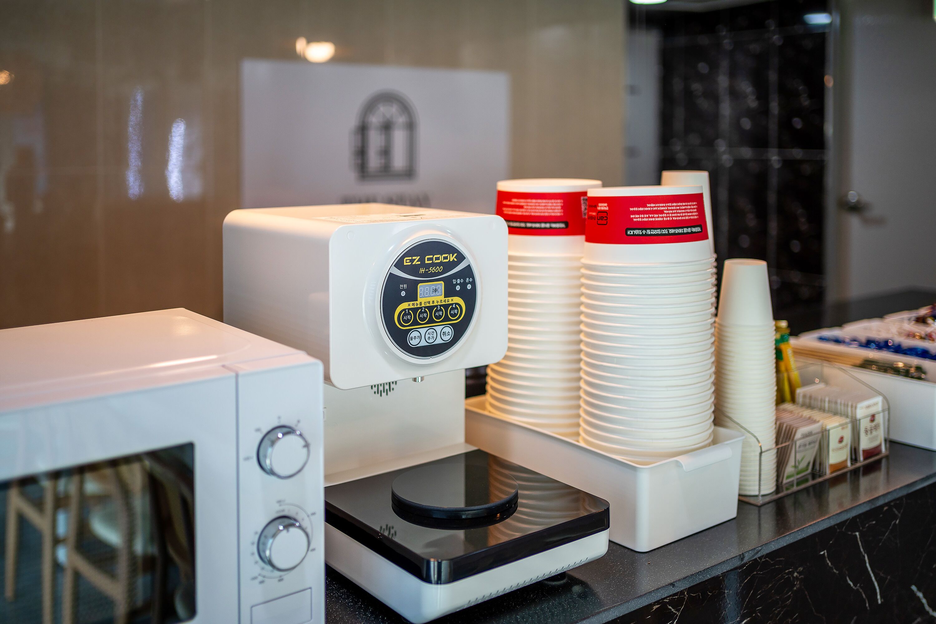 A coffee machine with a digital display, stacked white cups, and a tray of condiments on a hotel counter.
