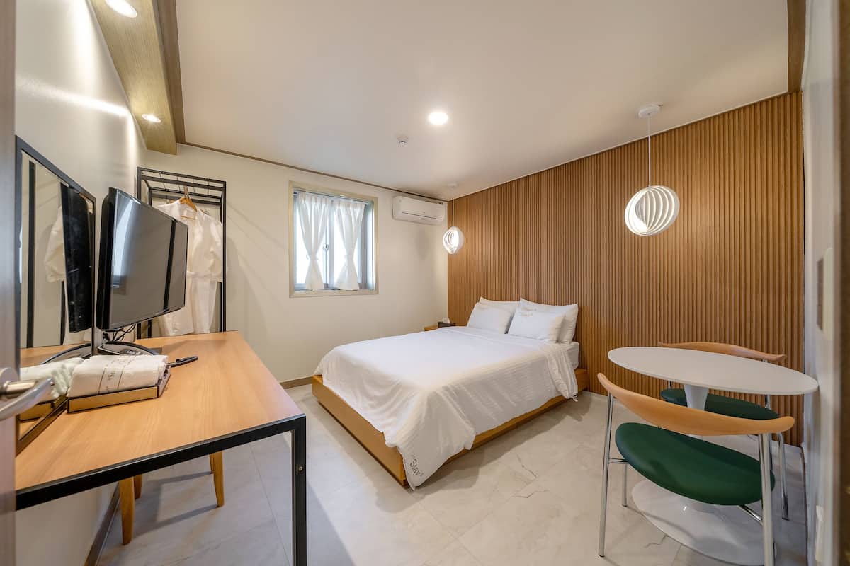 A hotel room with a bed, a desk with a computer, a chair, a small table, and a window with curtains.