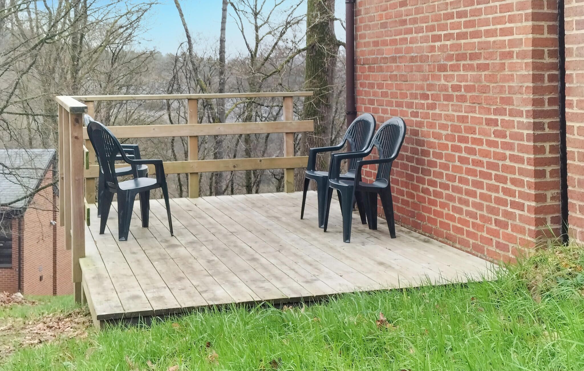 Une terrasse en bois avec deux chaises en plastique noir et une petite table, située à côté d’un mur en briques.