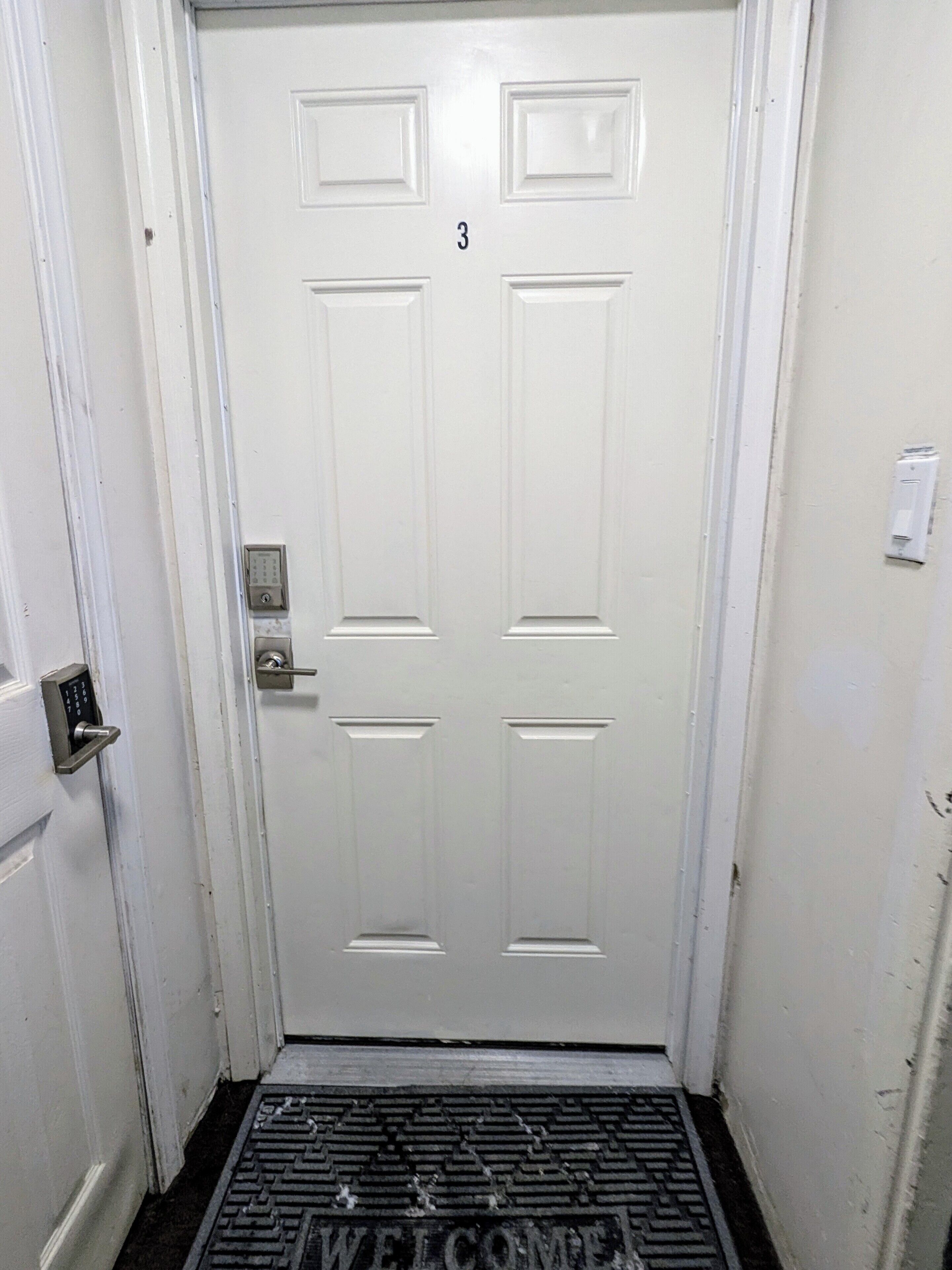 Una porta bianca con il numero '3' sopra, un campanello e un tappetino d'ingresso con la scritta 'WELCOME'.