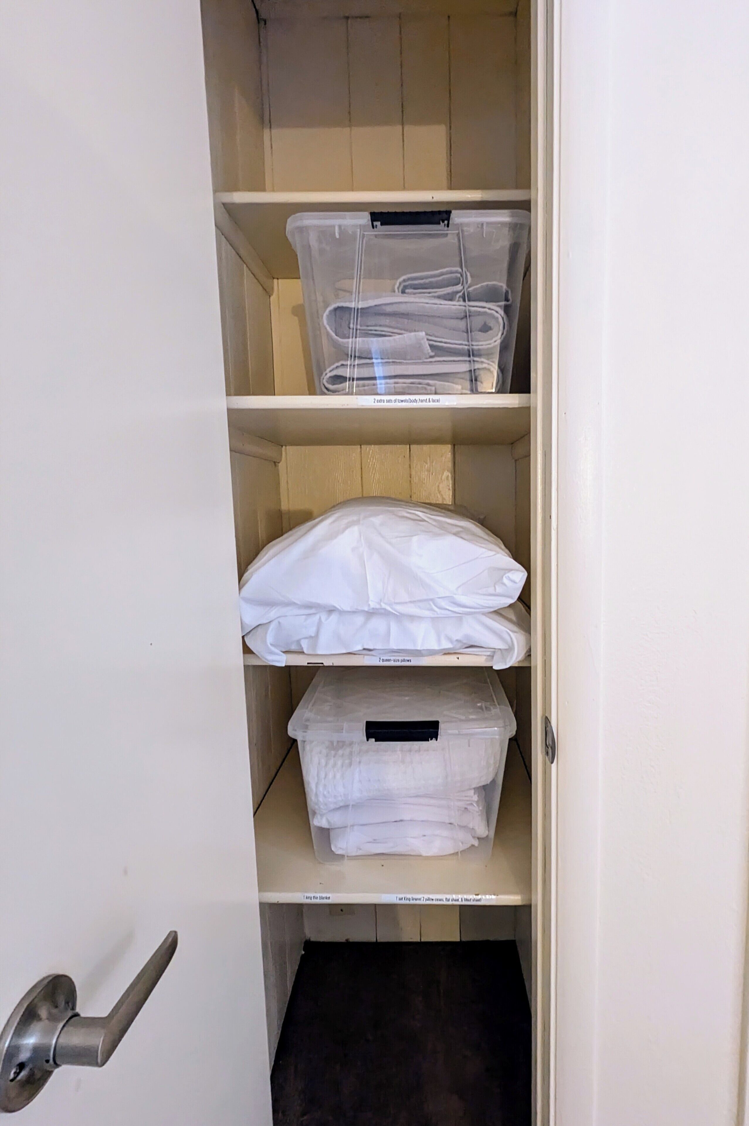 Et skab med hylder, der indeholder plastikbeholdere og foldet hvidt sengetøj.