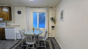 Una cocina compacta con comedor, que incluye una mesa redonda de vidrio, sillas blancas, un reloj y una ventana con cortinas.