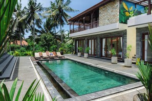 A modern pool area with lounge chairs, a stone building with a balcony, and lush tropical vegetation. - Kubu Manah Ubud (Ubud)