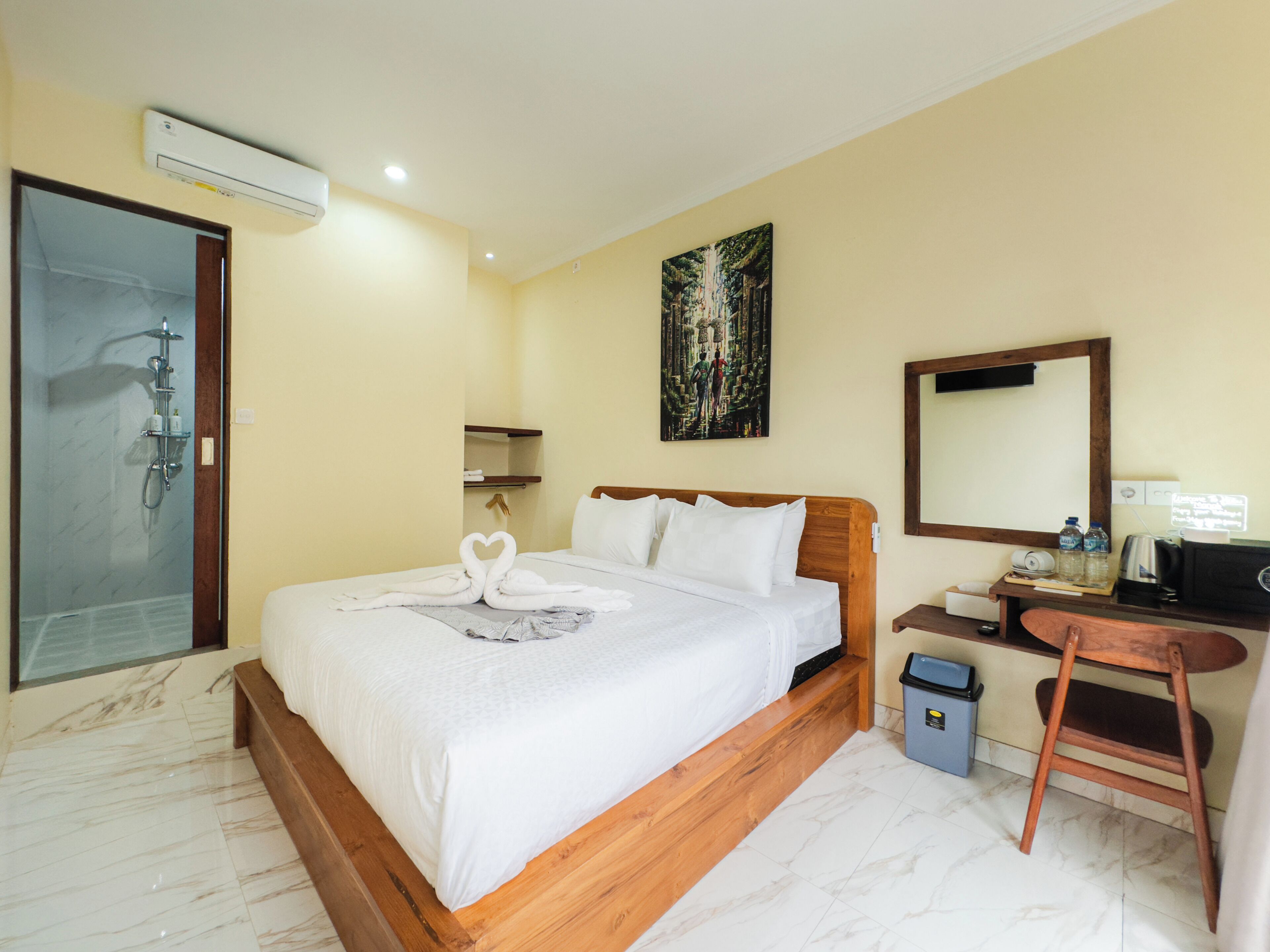 A hotel room with a bed, a shower, a desk with a chair, a trash bin, and a painting on the wall.