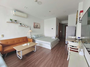Une pièce moderne et bien éclairée, avec un canapé, un lit, une petite table et un bureau doté d’un miroir. Des étagères murales, ornées de plantes et d’un tableau, complètent la décoration.