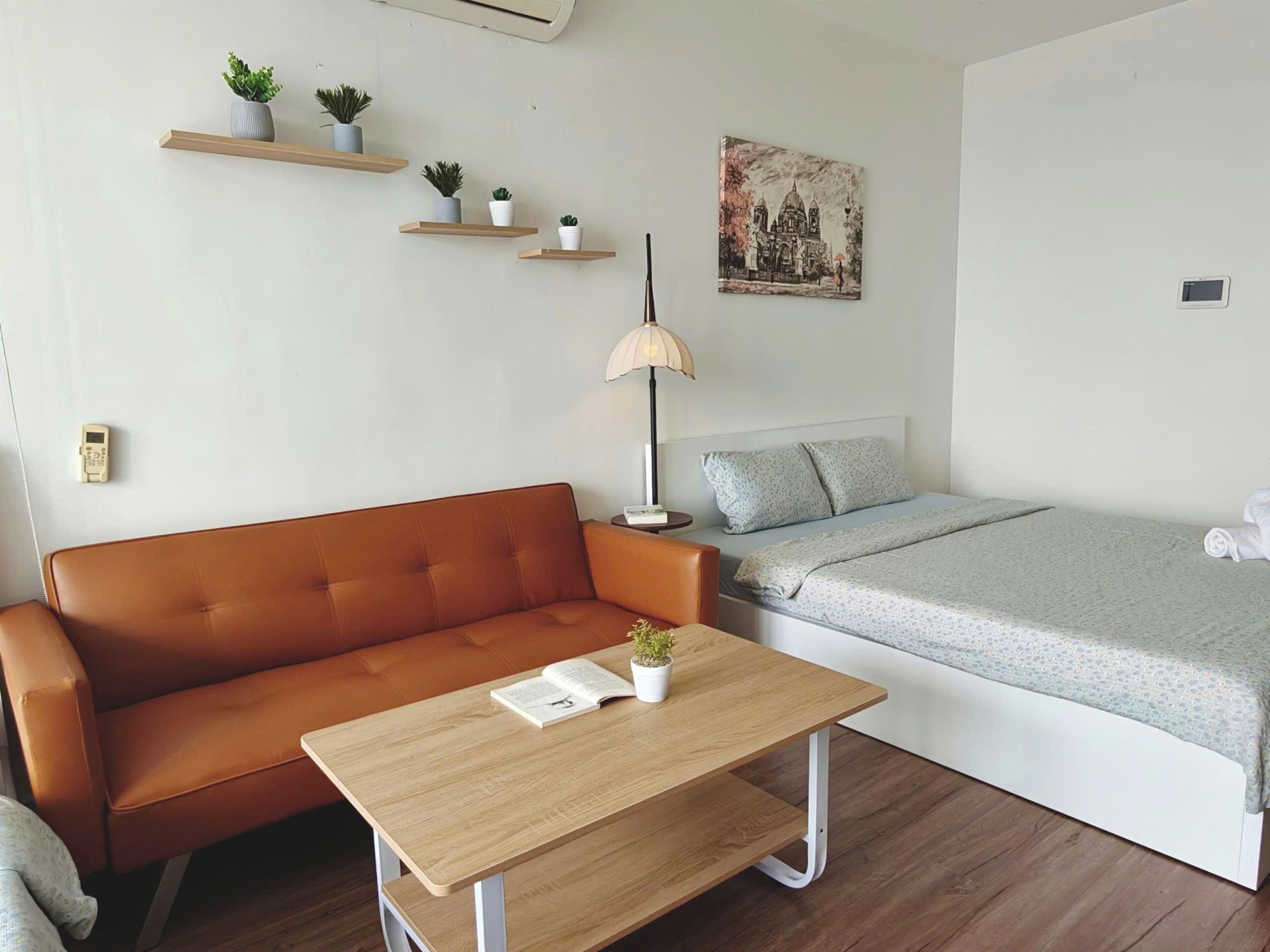 Une pièce avec un canapé marron, une table en bois sur laquelle se trouvent un livre et une plante, un lit recouvert d’une couverture grise et un tableau accroché au mur.