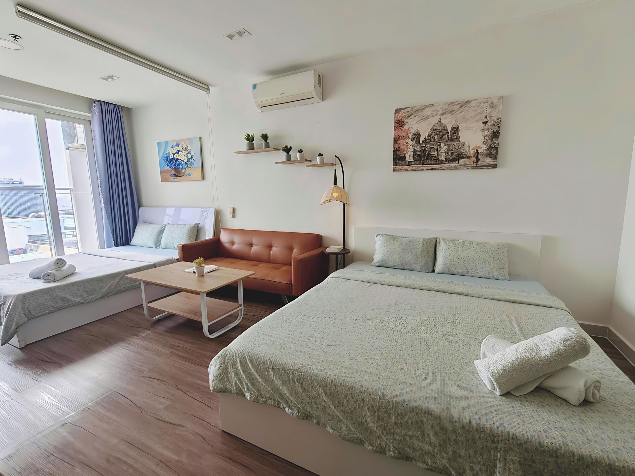 Habitació d'hotel amb llit, sofà, taula petita, finestra amb cortines, aire condicionat, prestatge fixat a la paret amb plantes i un quadre.