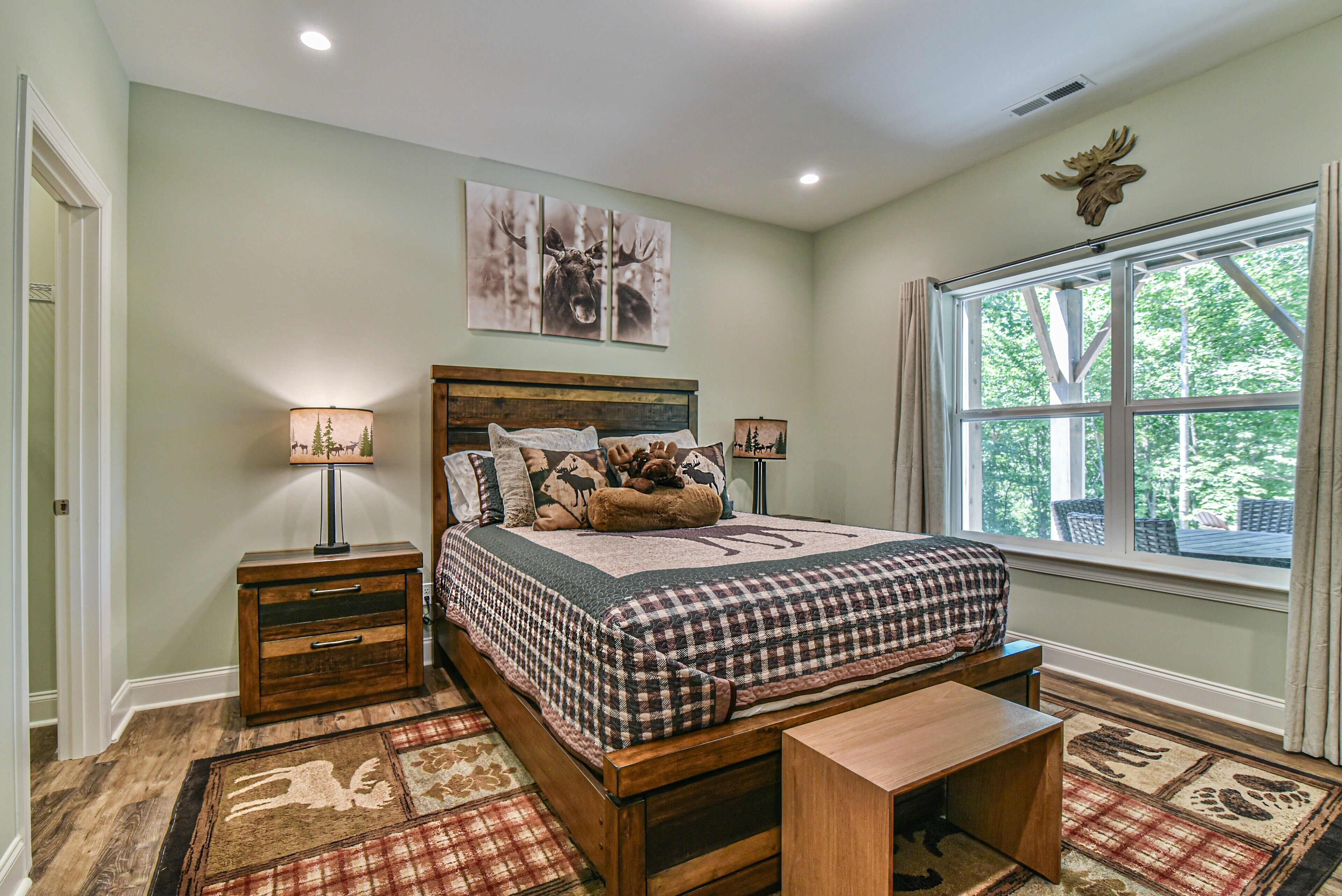 A bedroom with a wooden bed, two bedside tables, a bench, a window with a view of trees, and two wall-mounted lamps.
