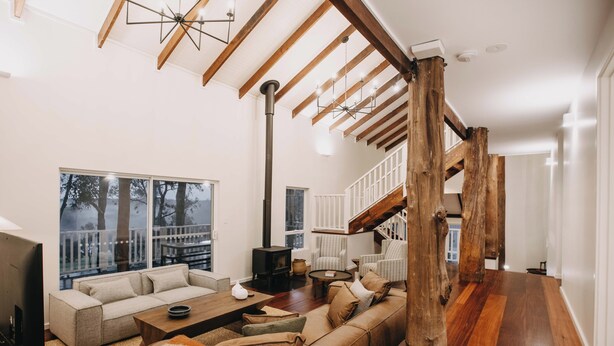 A spacious living room with a large sofa, a coffee table, and a fireplace. There is a staircase with wooden beams and a view of the outside through large windows. - The Vintners: Where luxury meets nature, five master suites & spacious living (Peerabeelup)