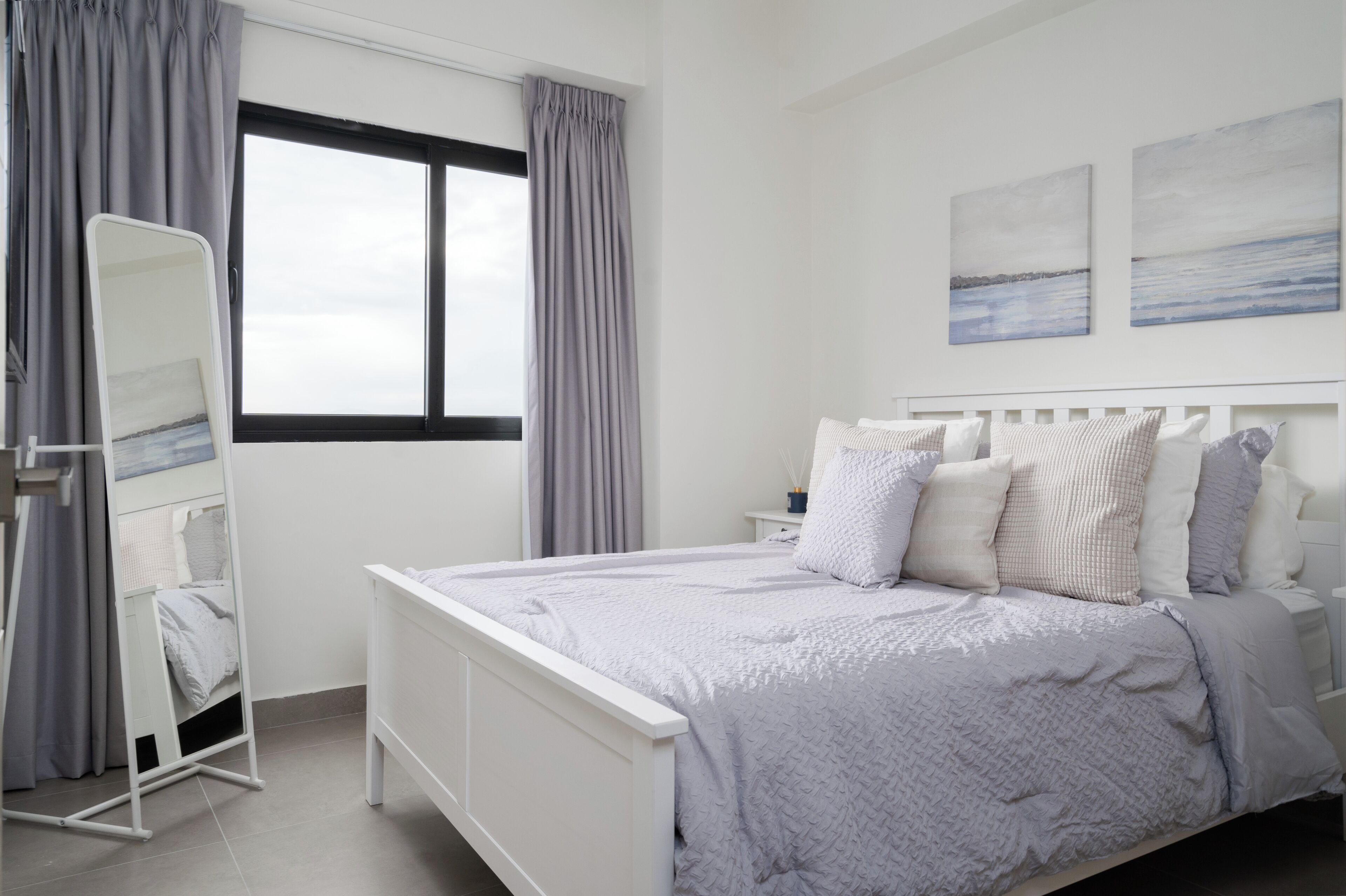 A neatly arranged bedroom with a bed, a bedside table, a mirror, and a window with curtains.