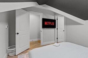 Ein Schlafzimmer mit einem Bett, einer weißen Wand, einer Tür und einem Fernseher, der 'NETFLIX' anzeigt.