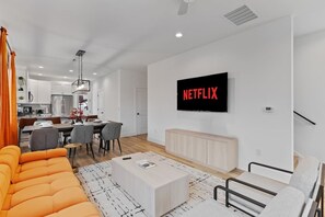 Una sala moderna con un televisor de pantalla plana mostrando 'NETFLIX' en la pared, un comedor con una cocina al fondo y un sofá naranja brillante.