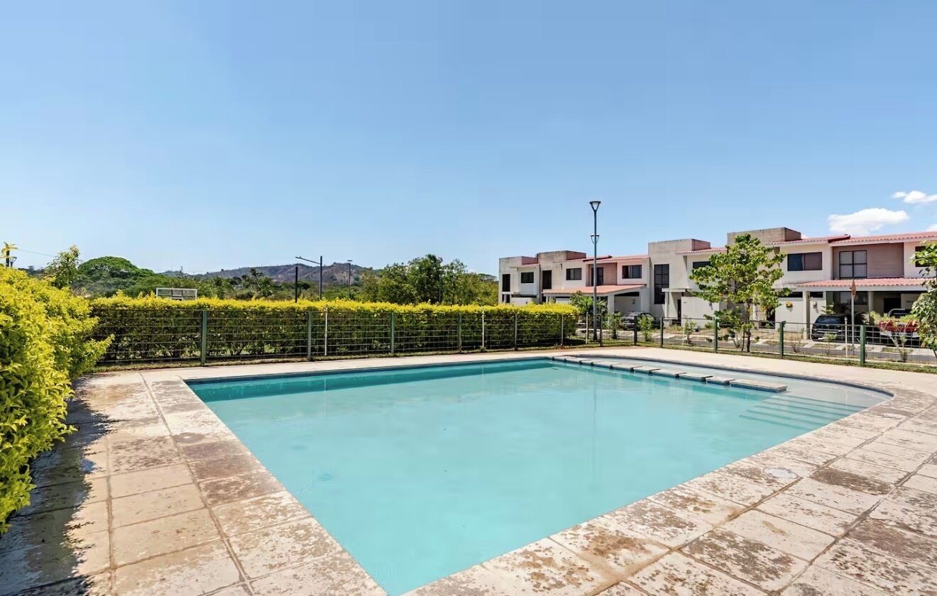 Una piscina rectangular con borde de azulejos, rodeada por un seto bajo y una zona pavimentada.