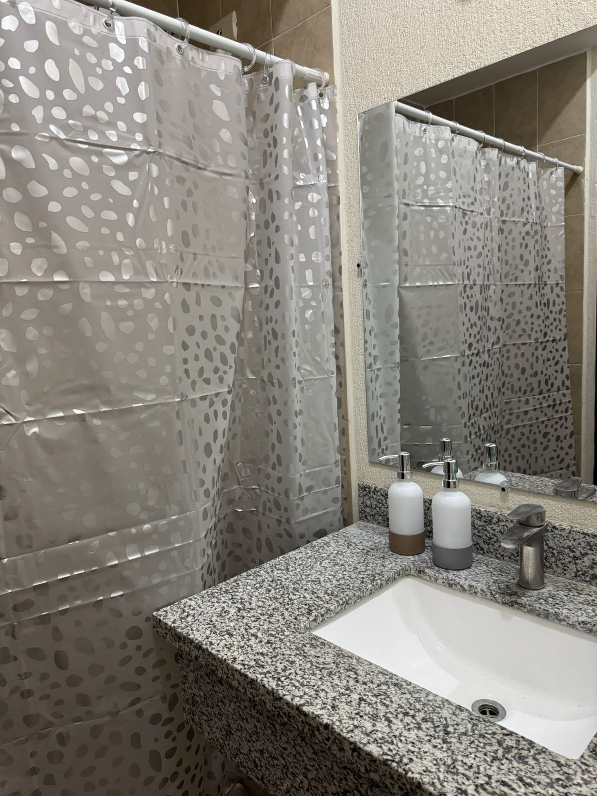 Un baño con encimera de granito, un lavabo blanco, dos dispensadores de jabón y una cortina de ducha con diseño de hojas.