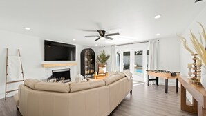 Una sala de estar moderna con un sofá seccional color beige, una chimenea, un televisor de pantalla plana, un ventilador de techo y una mesa de fútbol.