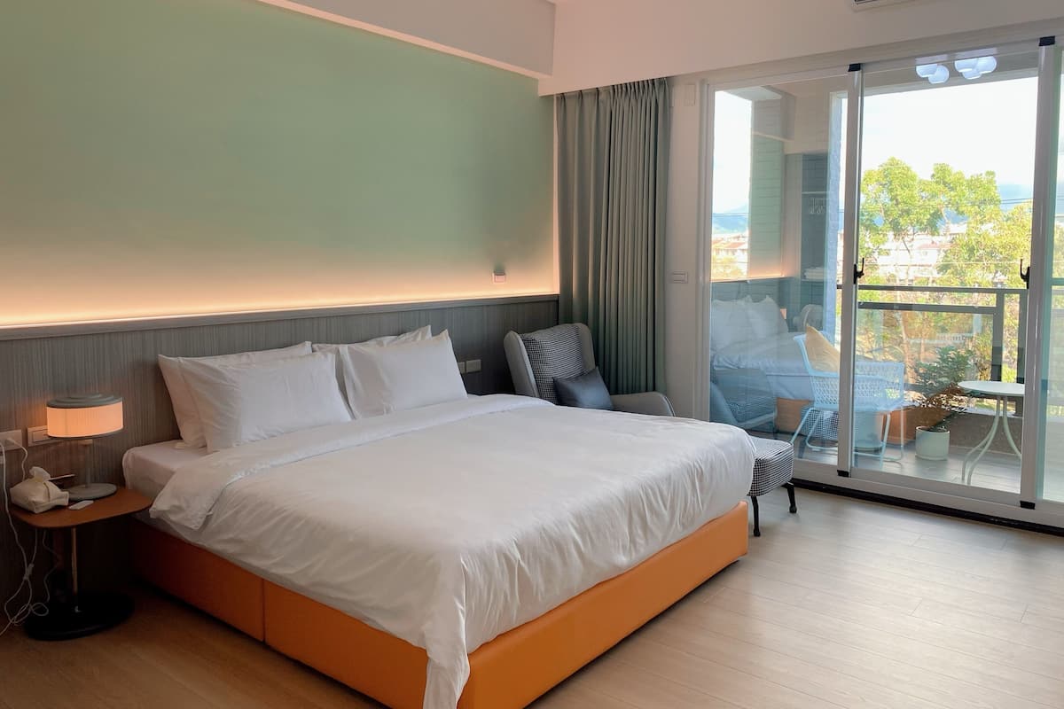 A hotel room with a large bed, a bedside table with a lamp, a chair, and a view of the outdoors through a sliding glass door.